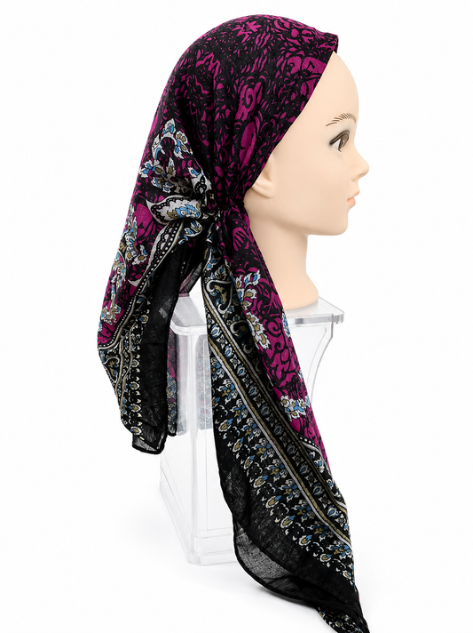 A patterned headscarf displayed on a mannequin head and clear stand against a white studio background. The scarf has a black base with a vivid magenta floral pattern and ornate multicolor border detailing.